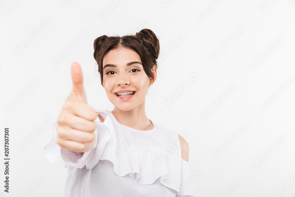 Portrait closeup of lovely student girl with double buns hairstyle and dental braces smiling while showing thumb up, isolated over white background Portrait closeup of lovely student girl with double buns hairstyle and dental braces smiling while showing thumb up, isolated over white background
