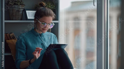Wallpaper Mural Stylish student girl sits by the window (on windowsill) with a gadget, listens to music. She is wearing a denim shirt, screen of the tablet is reflected in lens of glasses. Calm girl reads an e-book Torontodigital.ca