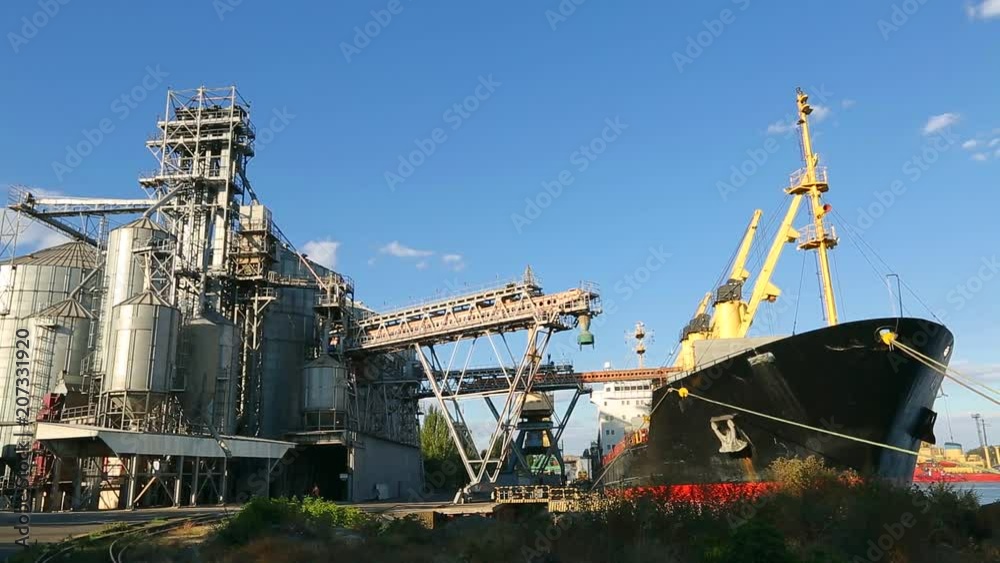 Panorama of grain terminal at seaport on sunny day. Cereals bulk transshipment to vessel loading grain crops on bulk ship from large elevators at the berth. Transportation of agricultural products. Panorama of grain terminal at seaport on sunny day. Cereals bulk transshipment to vessel loading grain crops on bulk ship from large elevators at the berth. Transportation of agricultural products.