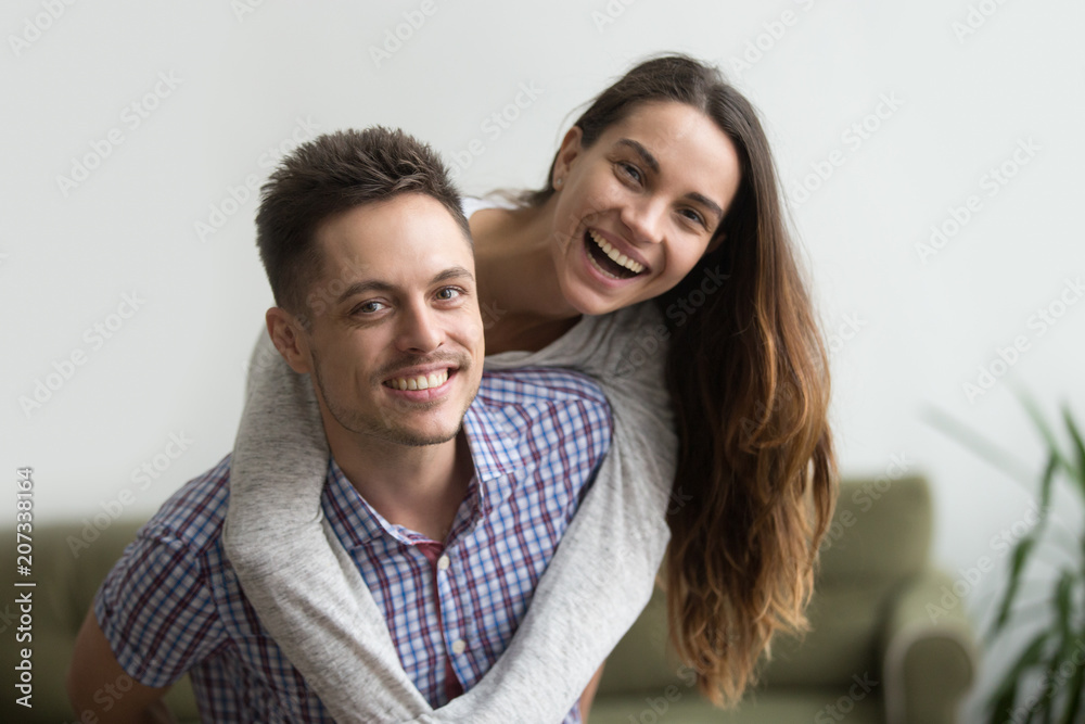 Obraz premium Smiling husband piggybacks cheerful wife at home, millennial couple in love embracing looking at camera, young real estate owners having fun in new house, happy man and woman dating headshot portrait