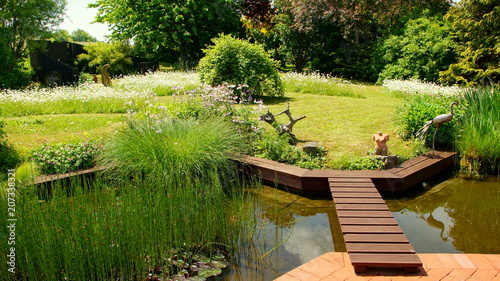 malerischer naturbelassener Garten mit Teich an roter Terrasse mit Holzbrücke und Blumeninseln