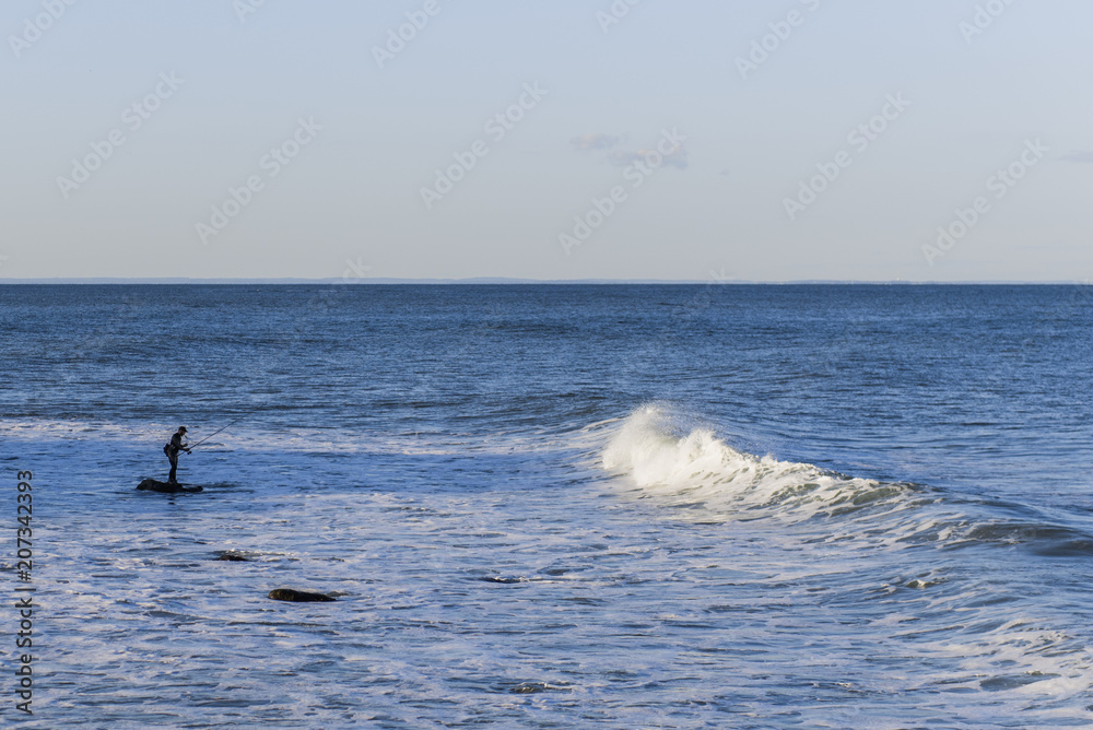 Fisherman in silhouette standing on a rock with wave rolling towards him.