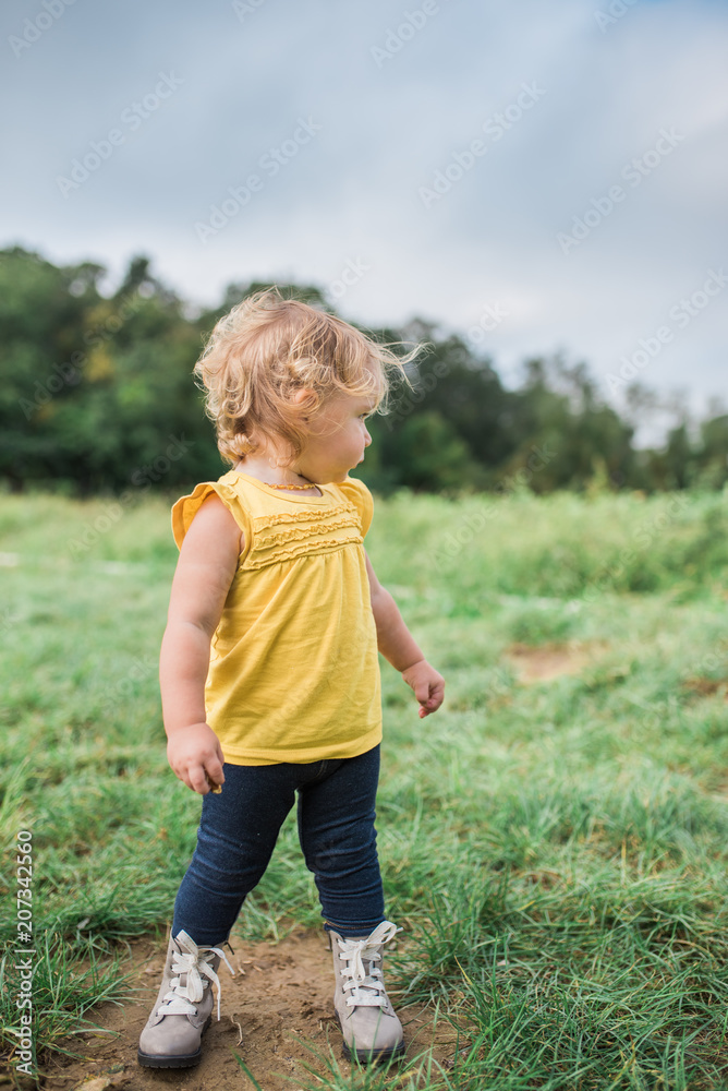 Blonde white female toddler wearing a yellow shirt, blue jeans and grey boots standing in a field and looking away from camera. Blonde white female toddler wearing a yellow shirt, blue jeans and grey boots standing in a field and looking away from camera.
