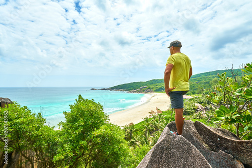 an adventure man hiking on neautifully shaped granite boulders and watching grand anse beach, la digue island, seychelles