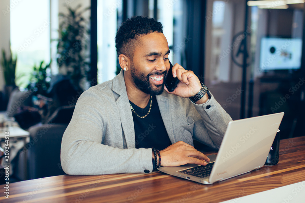 Smiling businessman working on laptop while talking on smartphone