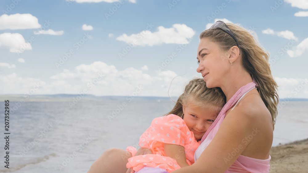 Little cute girl baby and young mother at beach. Mom sits on the shore, and her daughter is coming from behind. The kid hugs and kisses her mother. Mother and her child. Little cute girl baby and young mother at beach. Mom sits on the shore, and her daughter is coming from behind. The kid hugs and kisses her mother. Mother and her child.
