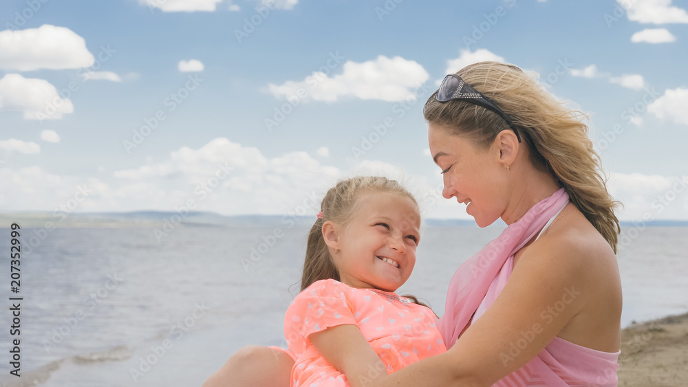 Little cute girl baby and young mother at beach. Mom sits on the shore, and her daughter is coming from behind. The kid hugs and kisses her mother. Mother and her child. Little cute girl baby and young mother at beach. Mom sits on the shore, and her daughter is coming from behind. The kid hugs and kisses her mother. Mother and her child.