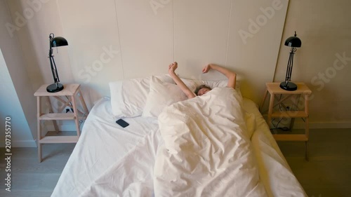Young Lady is Waking Up in a Cozy Bedroom and Turning Off the Alarm Clock on her Phone in the Sunny Morning photo