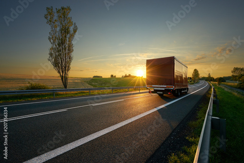 Obraz na płótnie Red small truck driving on asphalt road around farm fields in rural landscape at sunset w salonie