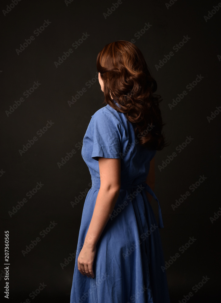 3/4 portrait of brunette lady wearing blue dress, facing away from camera. posed on black studio background.
