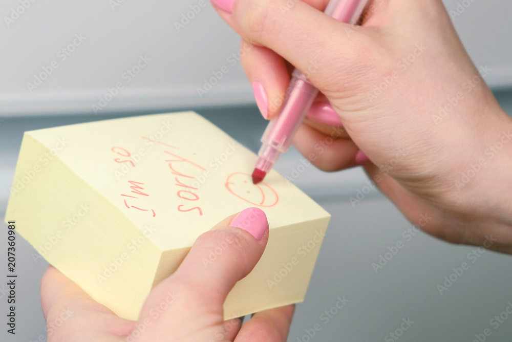 Closeup woman's hands write on yellow sticker paper sheets words i'm so sorry and laugthing emoticon. Closeup woman's hands write on yellow sticker paper sheets words i'm so sorry and laugthing emoticon.