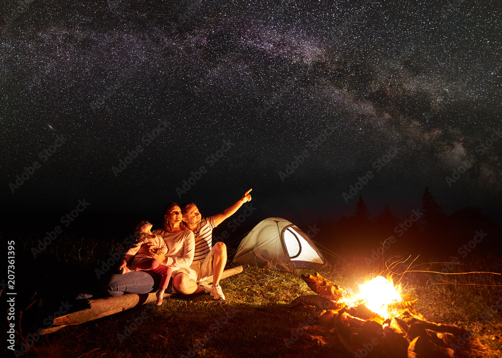Night camping in mountains. Family sitting in front of shining tent and burning campfire. Mother holding in arms small girl, father pointing at bright stars in dark sky. Tourism and traveling concept. - obrazy, fototapety, plakaty 