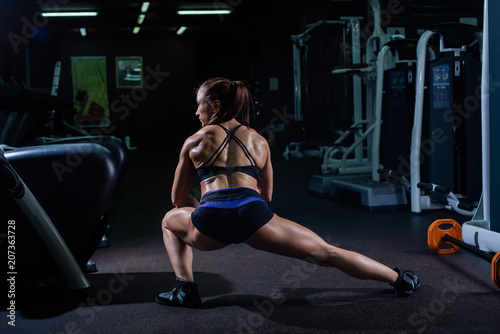 A strong, muscular, girl in black shorts, makes to strike forward and in side and stretches muscle legs. Dark gym.