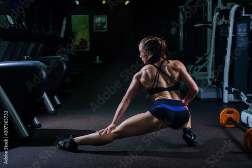 A strong, muscular, girl in black shorts, makes to strike forward and in side and stretches muscle legs. Dark gym.