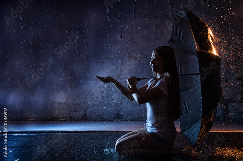 Girl in the white shirt with black umbrella, water drops around and dark walls background illuminated by light during a photoshoot with water