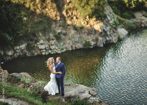 Beautiful and lovely newlyweds stand on a cliff, against the background of rocks and river. Wedding portrait of a stylish bride and a cute bride.