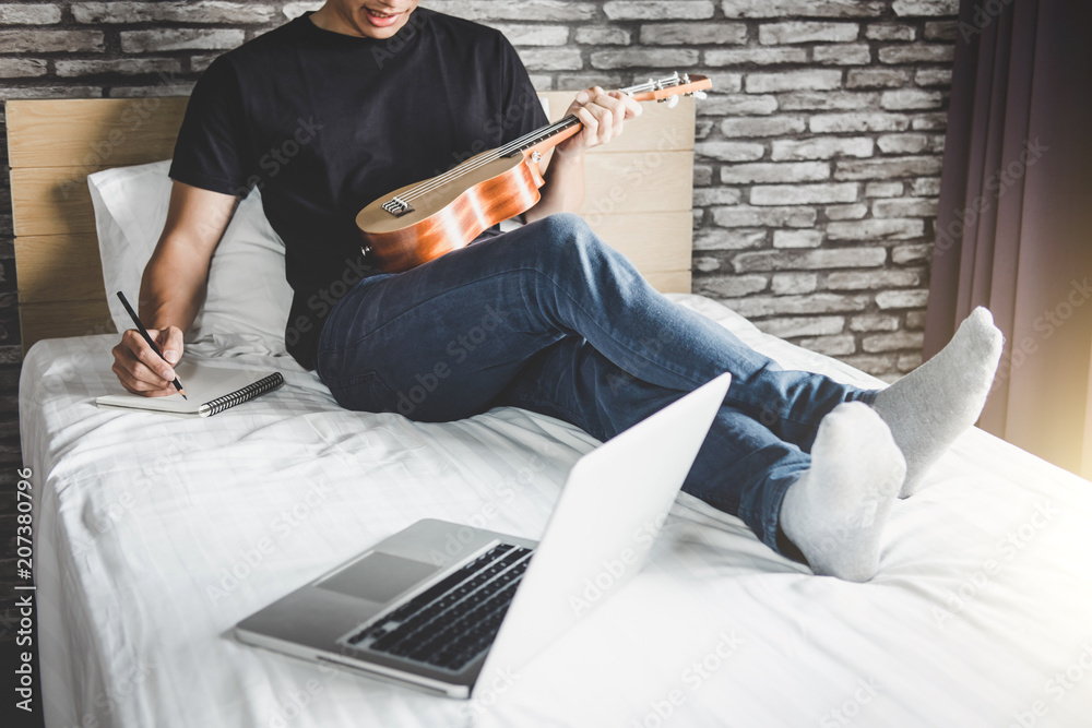 Young happiness man on bedroom in enjoying playing the music and compose a song with guitar or ukulele in holiday, relaxation and recreation concept