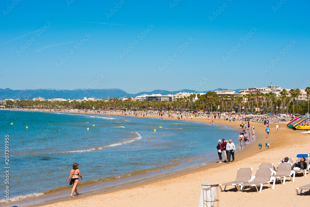 SALOU, TARRAGONA, SPAIN - APRIL 24, 2017: People walking along the beach of the Costa Dorada. Blue sky. Copy space for text. SALOU, TARRAGONA, SPAIN - APRIL 24, 2017: People walking along the beach of the Costa Dorada. Blue sky. Copy space for text.