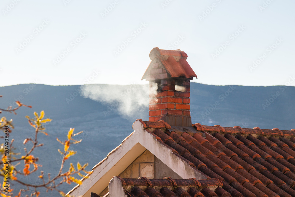 Stove heating. Smoke comes from brick chimney of the house with tiled roof. Country house near mountains in cold autumn.