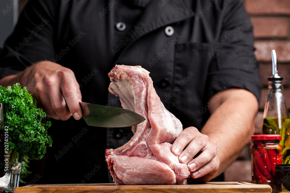 The concept of cooking meat. The man chef, the butcher, cuts raw meat with beef, lamb, veal, holding a knife in his hand, on a wooden table, next to lie raw vegetables, cherry, parsley, dill.