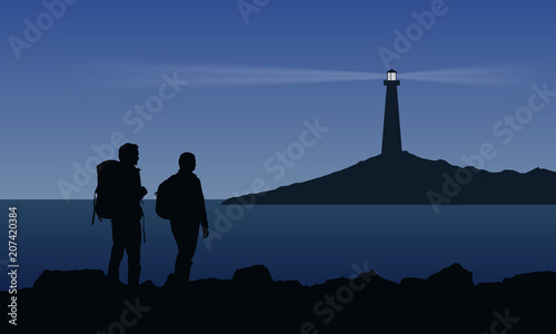 Two tourist with backpacks standing on the sea shore and watching the shining lighthouse on the island under blue night sky