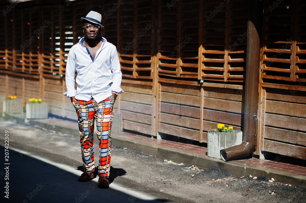 Stylish african american man in white shirt and colored pants with hat and glasses posed outdoor. Black fashionable model boy.