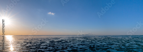 It is evening, the sun is aproaching the horizon and there is hardly any water in the Wadden Sea, it is low tide. On the horizon industrial facilities are visible - photographed by Greetsiel, Germany