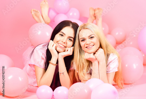Brunette and blonde cute women in pink pajamas posing on camera while lay near air balloons at slumber party over pink background