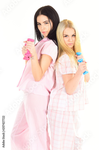 Morning exercises concept. Blonde and brunette on serious faces care about figure, doing exercises. Girls holds dumbbells, isolated on white background. Sisters or best friends in pajamas do sport.