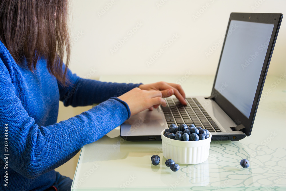 Fototapeta premium Young woman using laptop computer at home and eating blueberry.