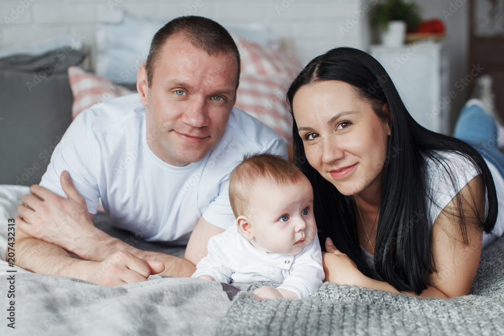 Obraz premium Young mom and dad and their baby son lie in bed and look at the camera indoors. The concept of a happy family