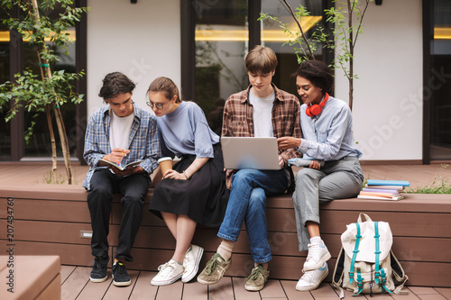 Group of students sitting on bench using laptop and reading book while spending time in courtyard of university. Company of young students study together outdoors