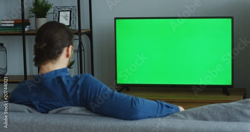 View from behind of the man in the jeans blue shirt sitting on the couch in the living room and watching TV with green screen. Chroma key. Indoors