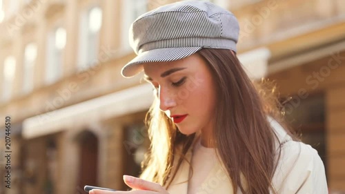 beautiful young woman with red lips standing against of bright sunshine on the street and using smartphone looking at screen thoughtful calm face blurred city life on background modern lifestyle photo