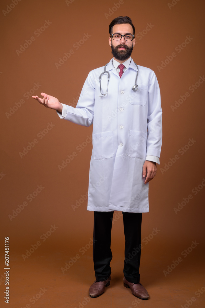 Young handsome bearded Persian man doctor against brown backgrou Young handsome bearded Persian man doctor against brown backgrou