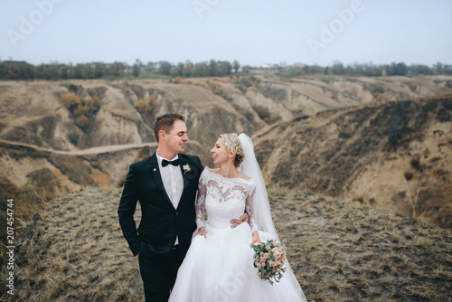 Tableau sur toile A stylish bridegroom in a black suit and a beautiful bride in a lace dress go and smile on the background of gray rocks and hills