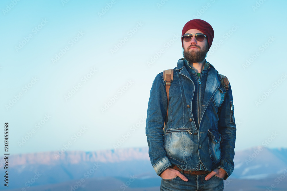 Fashion portrait of a bearded hipster young man wearing sunglasses, a backpack and hat on a background with copyspase in the mountains at sunset .. A confident man with a beard. Fashion portrait of a bearded hipster young man wearing sunglasses, a backpack and hat on a background with copyspase in the mountains at sunset .. A confident man with a beard.