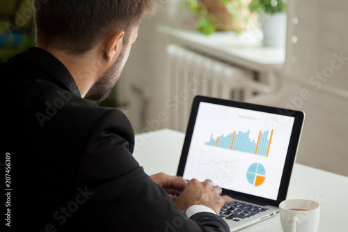 Fototapeta Naklejka Na Ścianę i Meble -  Thoughtful businessman working at laptop with financial data, analyzing growing rates and sales, planning future business strategy, preparing annual report. Motivation, leadership concept. Back view