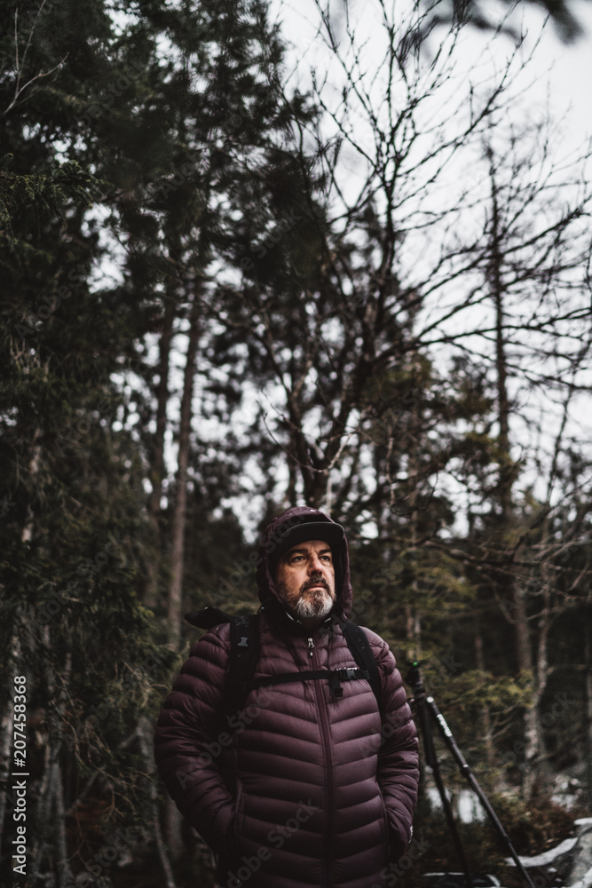 Älterer Mann mit grau melierten Vollbart steht mit Jacke und Kaputze und Rucksack auf dem Rücken im Wald und schaut in die ferne Landschaft Älterer Mann mit grau melierten Vollbart steht mit Jacke und Kaputze und Rucksack auf dem Rücken im Wald und schaut in die ferne Landschaft