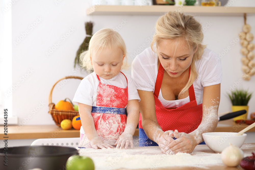 Little girl and her blonde mom in red aprons playing and laughing while kneading the dough in the kitchen. Homemade pastry for bread, pizza or bake cookies Little girl and her blonde mom in red aprons playing and laughing while kneading the dough in the kitchen. Homemade pastry for bread, pizza or bake cookies