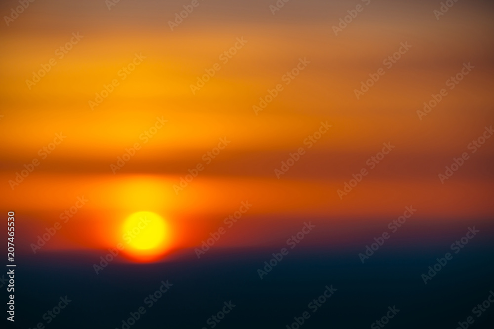 Blurry background image of circle of sun rising from behind dark horizon on background varicolored clouds of warmly shades. Abstract picturesque image of sun in bokeh.