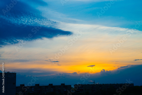 Atmospheric blue cloudy sky behind silhouettes of city buildings. Cobalt and orange background of sunrise with dense clouds and bright yellow sunny light for copy space. Cyan heaven above clouds.