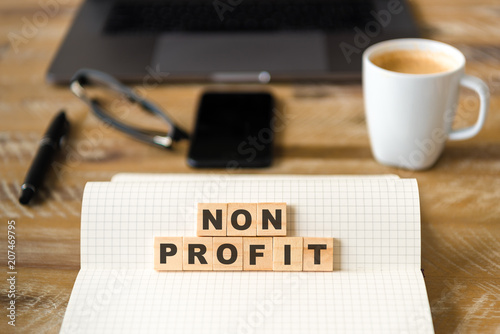 Closeup on notebook over wood table background, focus on wooden blocks with letters making Non Profit text