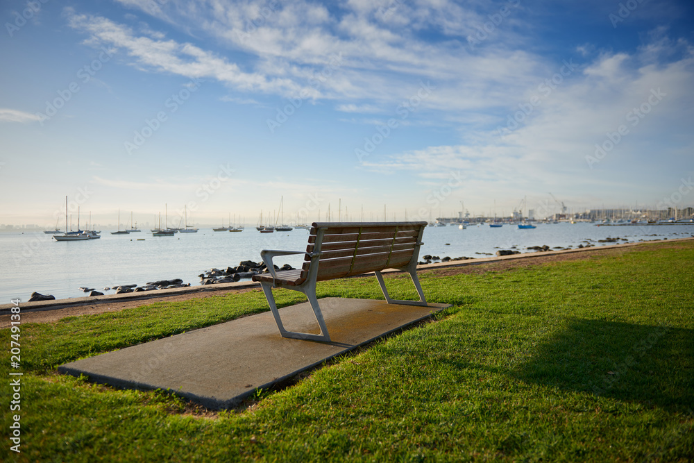 An empty bench on grass by the waters edge in the inspiring bright morning sun with a gorgeous view of boats and marina in the distance