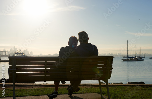 A silhouette of a couple sitting on a bench by the waters edge enjoying the sun in their face and vista of water and boats