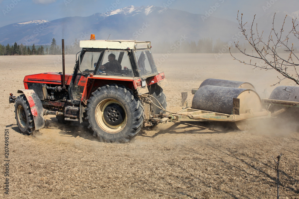 Obraz premium Tractor turning when pulling heavy metal roller, preparing dry field in the spring, dust cloud behind, with mountains in the background. (trademarks/face blurred/cloned)