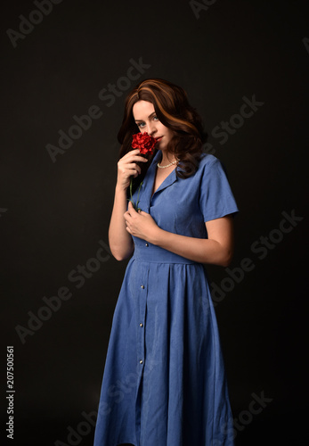 3/4 portrait of brunette lady wearing blue dress holding a red flower. posed on black studio background.