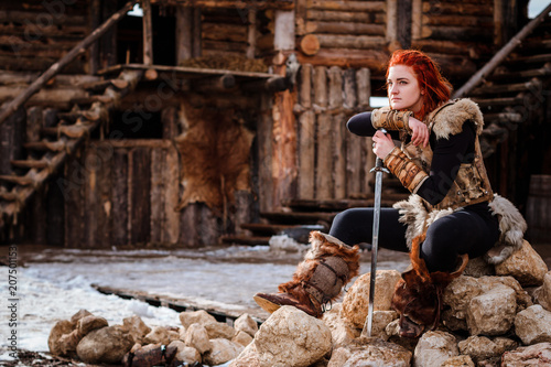 Red-haired woman is a Viking. Against the backdrop of a village or an ancient settlement. In the hands of a sword photo