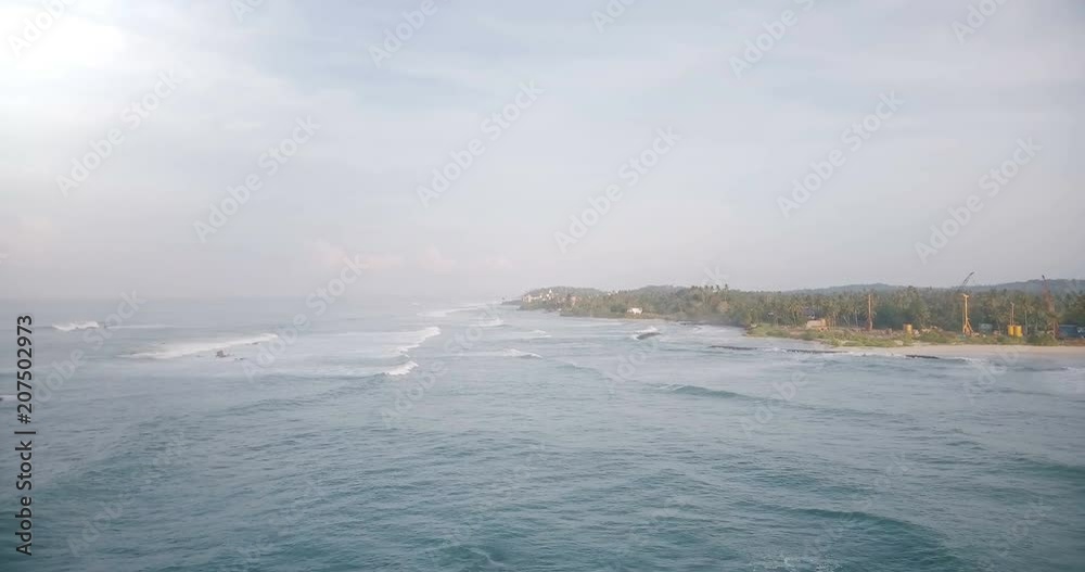 Aerial drone shot of beautiful foaming sea waves reaching tropical shore with green trees, houses and construction site. Aerial drone shot of beautiful foaming sea waves reaching tropical shore with green trees, houses and construction site.