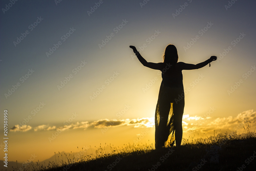 silhouete of girl in dress standing on grass in sunset mountains